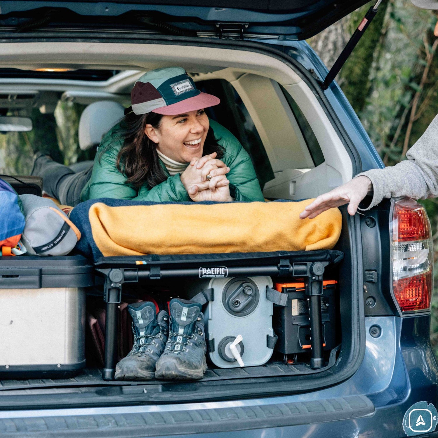 Person sitting in an open car trunk with camping gear, including a yellow sleeping pad and boots.